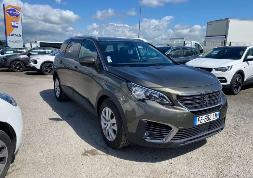 Peugeot 5008 gris clair vue 3/4 avant droit sur parking avec ciel partiellement nuageux.