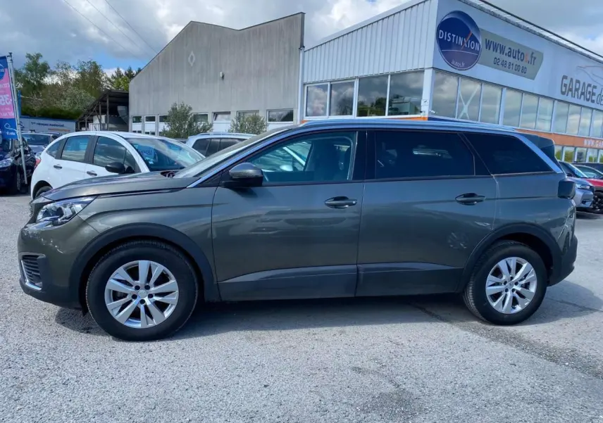 Profil côté gauche d'un Peugeot 5008 gris clair de 2019 garé devant un garage avec vitres surteintées.