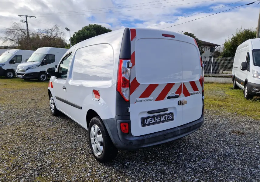 Vue 3/4 arrière droite d’un Renault Kangoo blanc avec bandes rouges réfléchissantes sur les portes arrière, stationné sur un sol gravillonné.