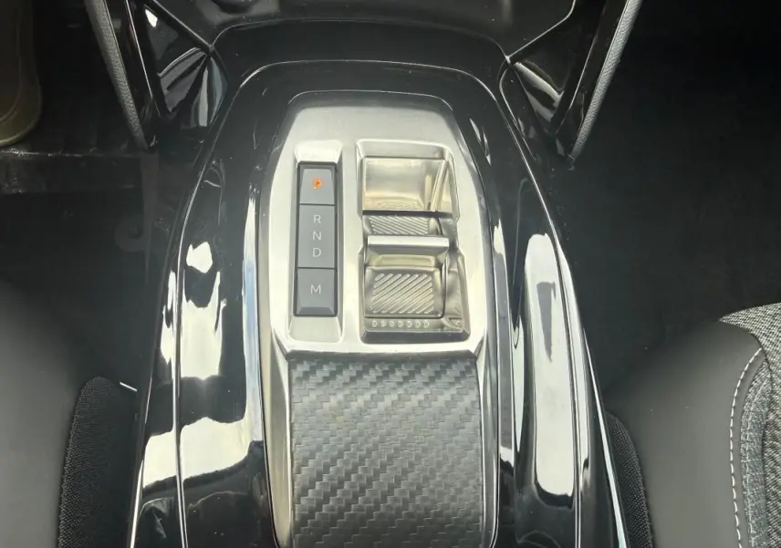 Vue plongeante sur la console centrale noire brillante de la Peugeot 208 Hybrid 145 avec levier de vitesses électronique et finition carbone.