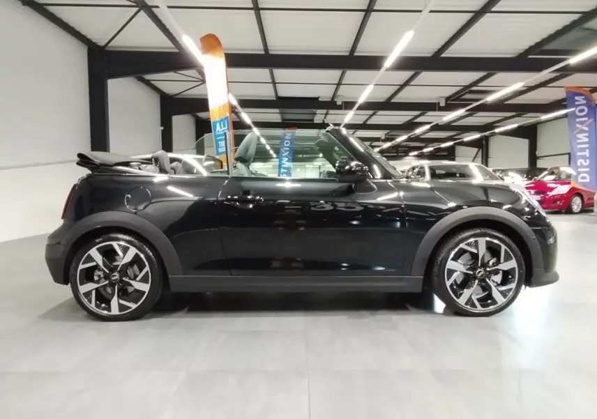 Profil droit d'une MINI Cabriolet noire 2025 avec capote ouverte et jantes 16 pouces 4-Square Spoke en intérieur showroom.