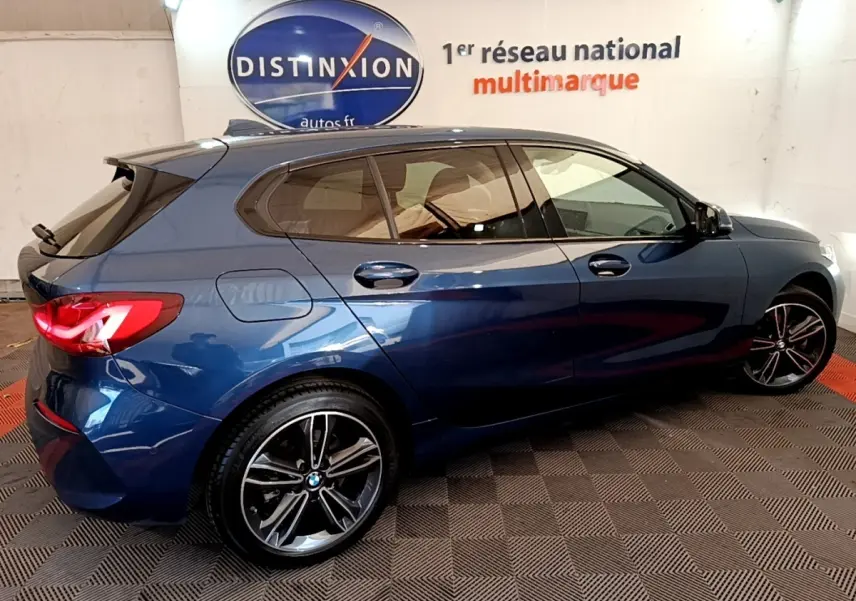 BMW Série 1 118i Edition Sport bleu vue de profil droit en intérieur, avec jantes alliage et feux arrière LED allumés.