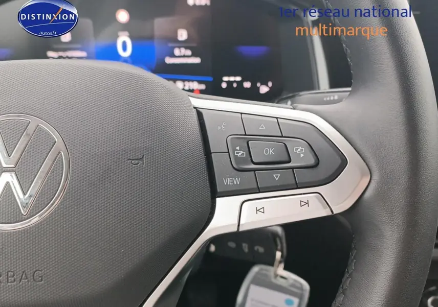 Gros plan sur le volant multifonction noir avec logo Volkswagen, tableau de bord digital flou en arrière-plan.