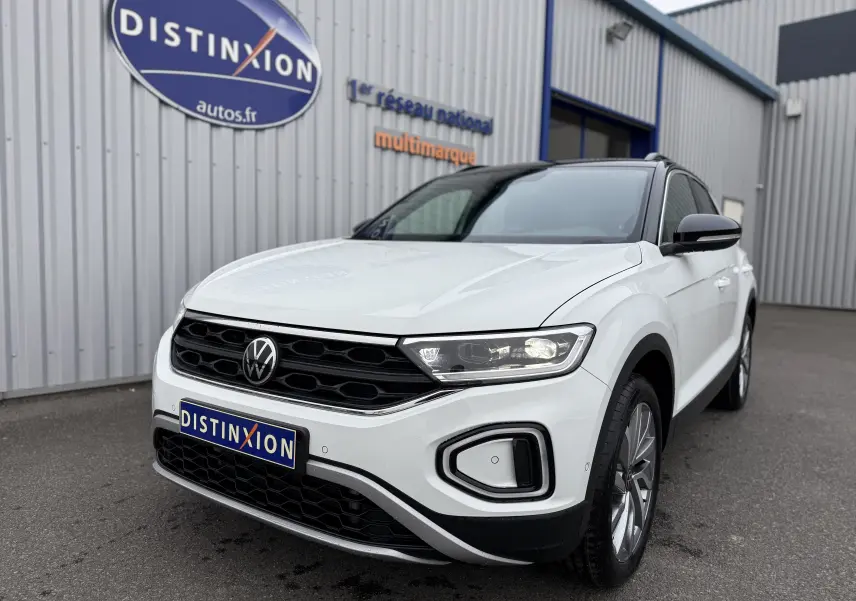 Volkswagen T-Roc blanc pure avec toit noir, vue 3/4 avant droit, phares LED allumés, devant un bâtiment gris.