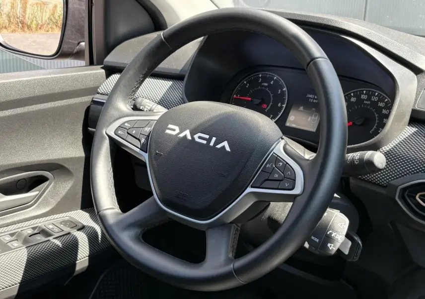 Vue intérieure centrée sur le volant noir Dacia avec tableau de bord et commandes au volant visibles.