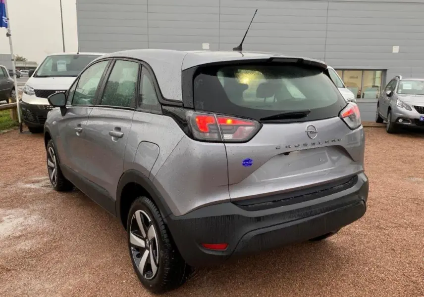 Vue 3/4 arrière droite d'un Opel Crossland gris métallisé avec feux arrière allumés sur parking humide.