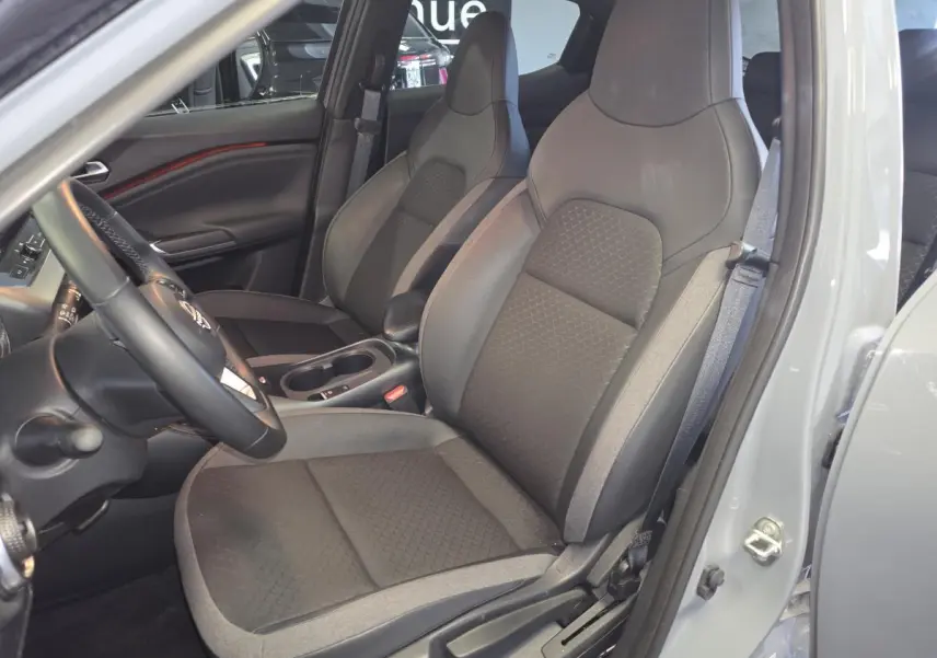 Vue intérieure côté conducteur du Nissan Juke 2025 gris, montrant les sièges avant en tissu et TEP noir et gris avec accoudoir central.