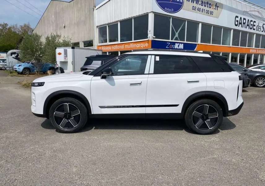 Vue de profil côté gauche du SUV JAECOO J7 blanc khaki avec toit noir et jantes modernes, stationné devant un garage.