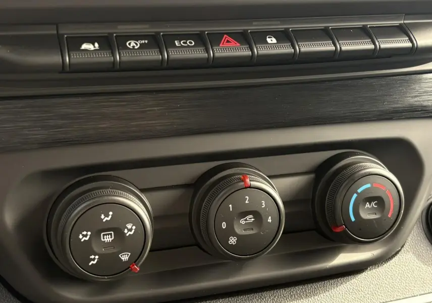 Gros plan sur les commandes de climatisation noires du Renault Trafic Fourgon blanc Glacier, avec habillage bois visible.