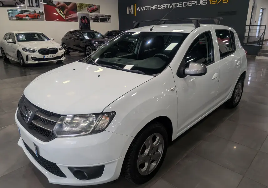 Vue 3/4 avant droite d'une Dacia Sandero TCe 90 blanche Glacier en showroom avec barres de toit noires.