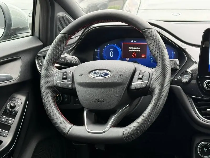 Vue rapprochée du volant perforé noir avec surpiqûres rouges du Ford Puma 2024, tableau de bord digital et commandes intégrées visibles.