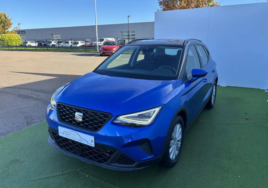 Vue 3/4 avant d'un SEAT Arona 2024 bleu saphir avec toit noir et jantes alliage, stationné sur sol vert.