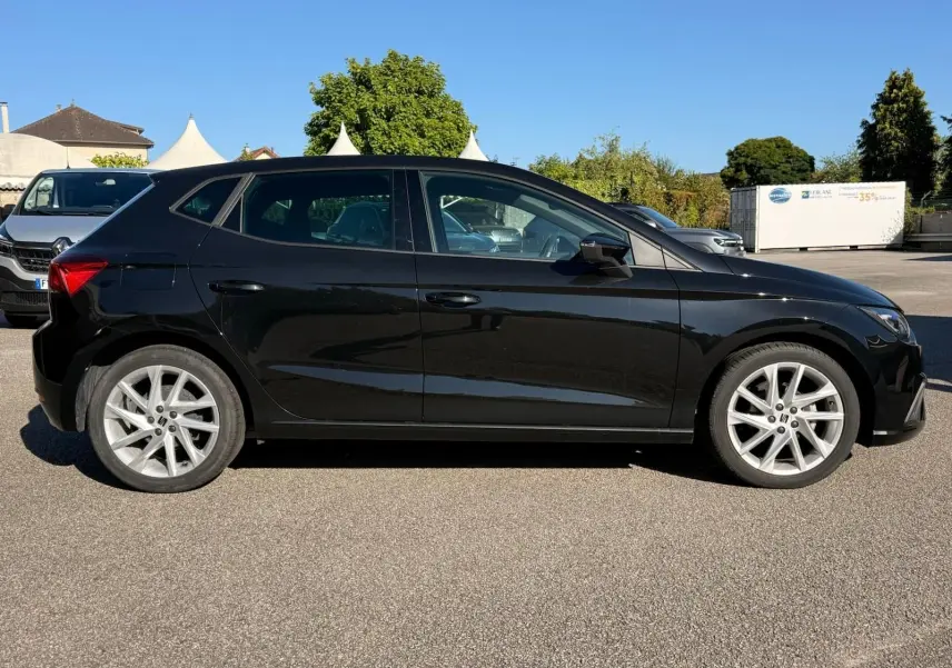 Profil droit d'une SEAT Ibiza 1.0 TSI 95 FR noire garée sur un sol goudronné sous un ciel bleu clair.