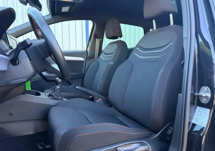 Vue intérieure côté conducteur de la SEAT IBIZA noire 2025, sièges tissu avec surpiqûres rouges et volant cuir.