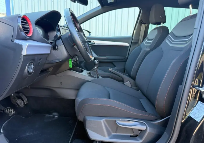 Vue intérieure côté conducteur de la SEAT Ibiza 1.0 TSI 95 FR noire, sièges tissu avec surpiqûres rouges et volant cuir.