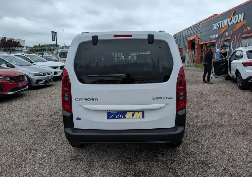 Vue arrière d'un Citroën Berlingo blanc avec hayon vitré et feux arrière rouges allongés, stationné sur un parking.