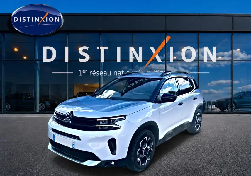 Citroën C5 Aircross blanc en 3/4 avant droit, avec jantes alu et barres de toit visibles devant un showroom.