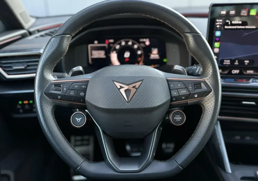 Vue rapprochée du volant cuir perforé noir avec logo CUPRA cuivré, tableau de bord digital et écran tactile en arrière-plan.