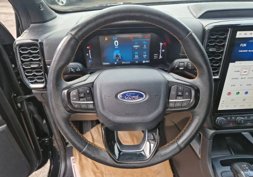 Vue intérieure centrée sur le volant noir en cuir avec surpiqûres orange du Ford Ranger Wildtrak 2023, tableau de bord digital visible.