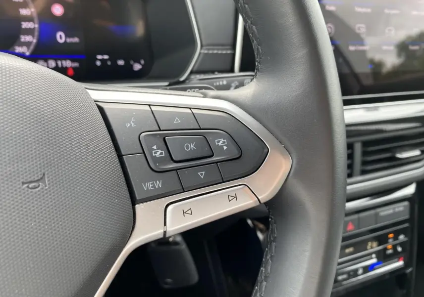 Gros plan sur les commandes gauche du volant cuir noir du Volkswagen T-Cross avec tableau de bord digital en arrière-plan.