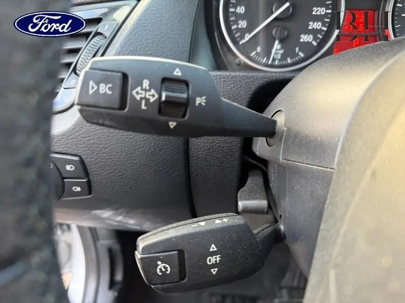 Gros plan sur les commandes au volant d'une BMW X1 Mineralgrau, avec levier de clignotant et régulateur de vitesse visibles.