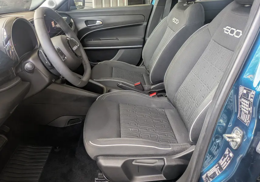 Vue intérieure côté conducteur de la FIAT 600 1.2 Hybrid 2025 avec sièges noirs marqués "600" et portière bleu Sea of Italy ouverte.