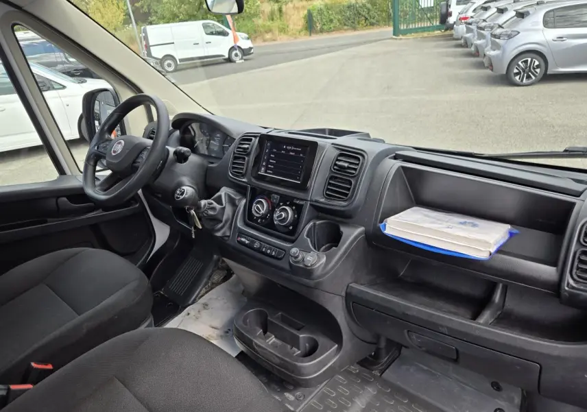 Vue intérieure côté conducteur du Fiat Ducato Fourgon blanc, montrant le tableau de bord avec écran tactile et volant noir.