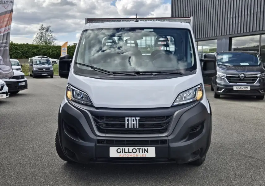 Vue frontale d’un FIAT Ducato Fourgon blanc 2022 avec calandre noire et phares allumés sur un parking extérieur.