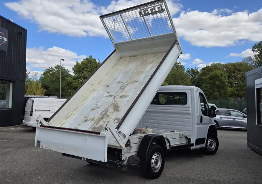 Fiat Ducato Fourgon blanc vu en 3/4 arrière avec benne basculante relevée sur parking extérieur.