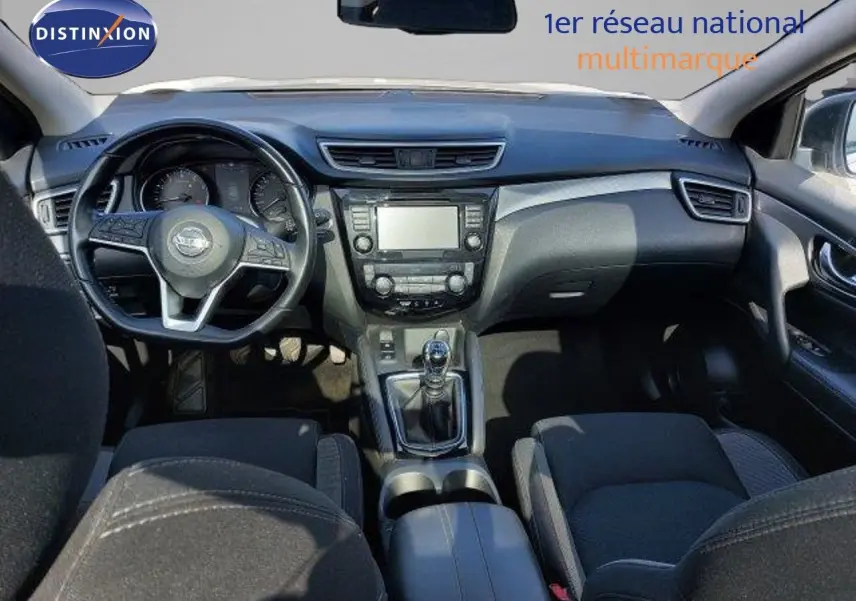 Vue intérieure du tableau de bord et console centrale du Nissan Qashqai blanc lunaire, avec volant et boîte manuelle visibles.