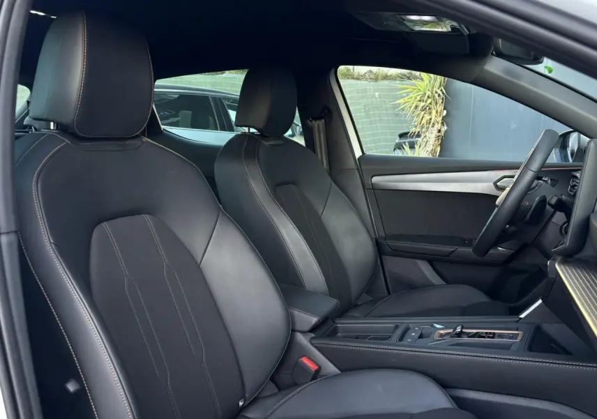 Vue intérieure côté conducteur de la CUPRA Leon 2025, sièges sports noirs avec surpiqûres contrastées et accoudoir central.