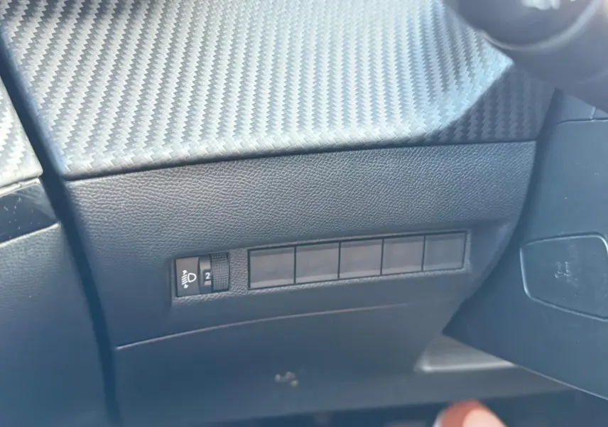 Détail intérieur côté gauche du tableau de bord noir avec commande de réglage des phares sur Peugeot 2008 Hybrid 136 e-DCS6 Allure.