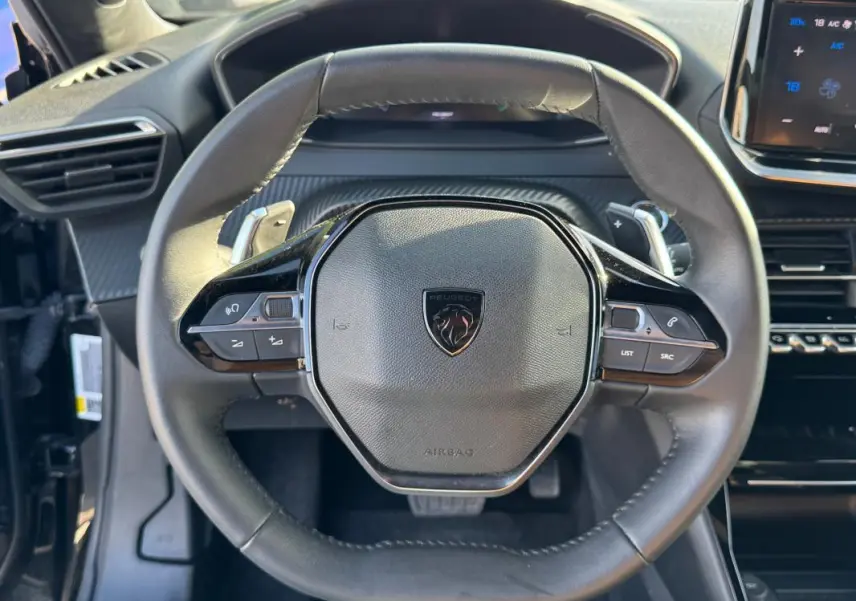 Vue rapprochée du volant noir de la Peugeot 2008 Hybrid 136 e-DCS6 Allure avec commandes intégrées et tableau de bord numérique.