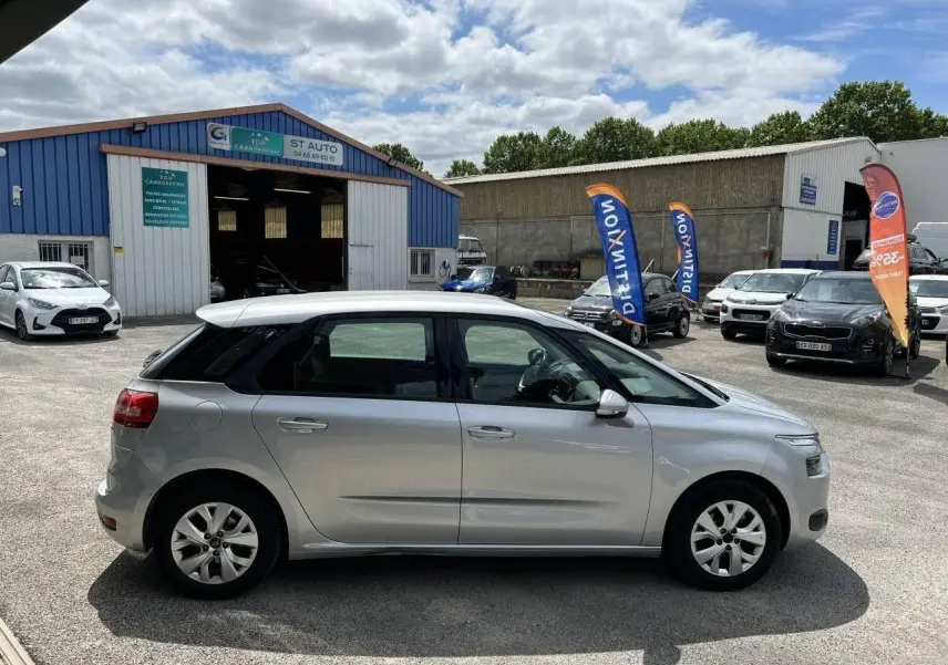 Profil côté droit d'une Citroën C4 Picasso gris aluminum 2016 stationnée devant un garage, sous un ciel partiellement nuageux.