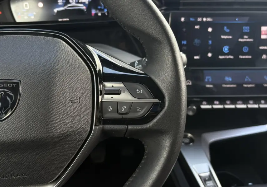 Gros plan sur le volant cuir noir de la Peugeot 308 2024 avec commandes vocales et écran tactile en arrière-plan flou.