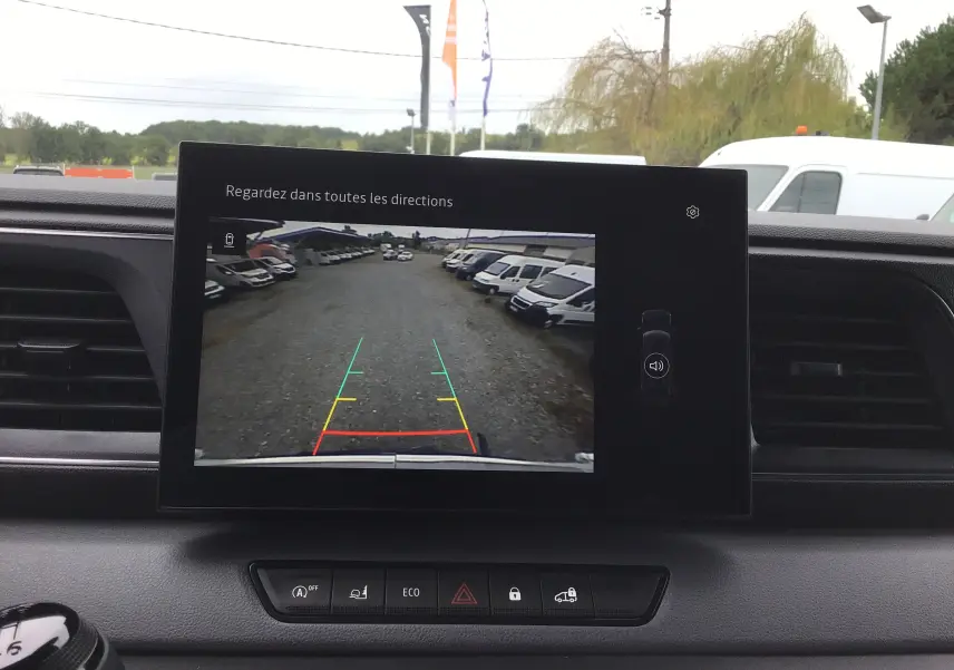 Écran multimédia du Renault Master blanc 2025 montrant la caméra de recul avec vue arrière sur un parking de véhicules blancs.