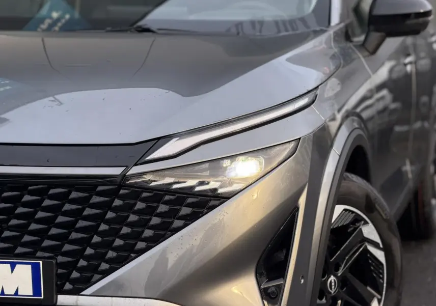 Gros plan sur l'avant droit du Nissan Qashqai 2025 gris Squale, avec phares allumés et jante alliage distinctive.