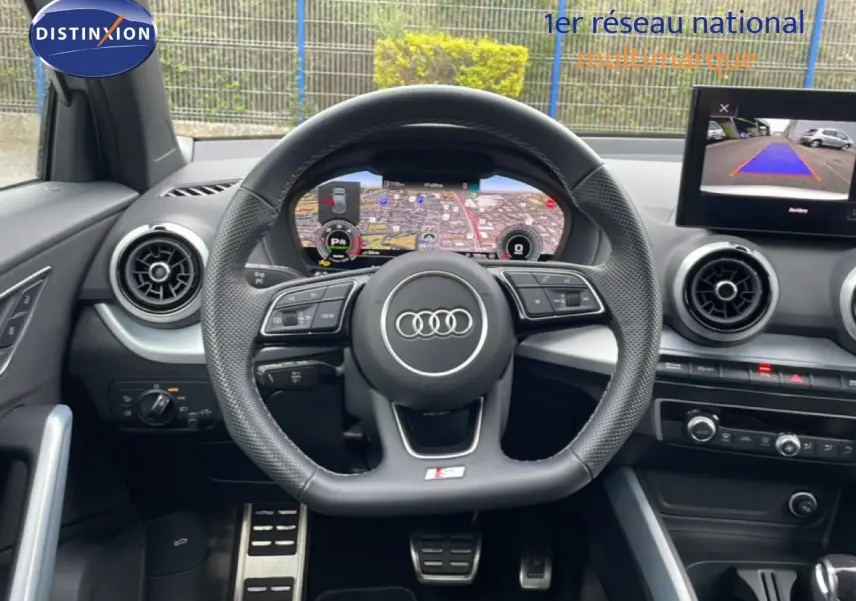 Intérieur Audi Q2 2025 vu du poste de conduite, volant cuir S-Line, tableau de bord numérique et écran caméra de recul.