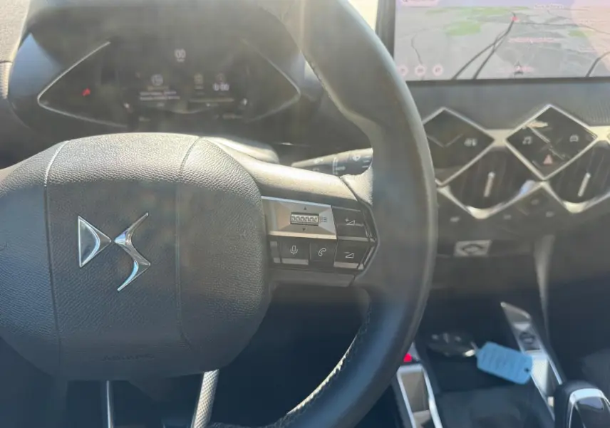 Vue rapprochée du volant DS d’une DS3 2024 gris, avec commandes intégrées et tableau de bord numérique en arrière-plan.