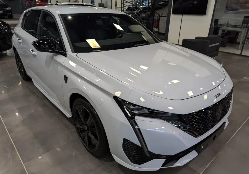 Peugeot 308 BlueHDi 130 GT blanc Okénite vue 3/4 avant droit avec jantes noires et calandre distinctive.