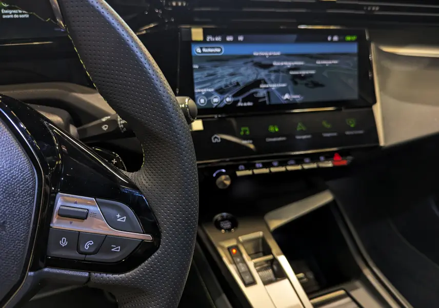 Vue rapprochée du volant et de l'écran tactile central de la Peugeot 308 GT blanche, mettant en valeur les commandes au volant et la navigation.
