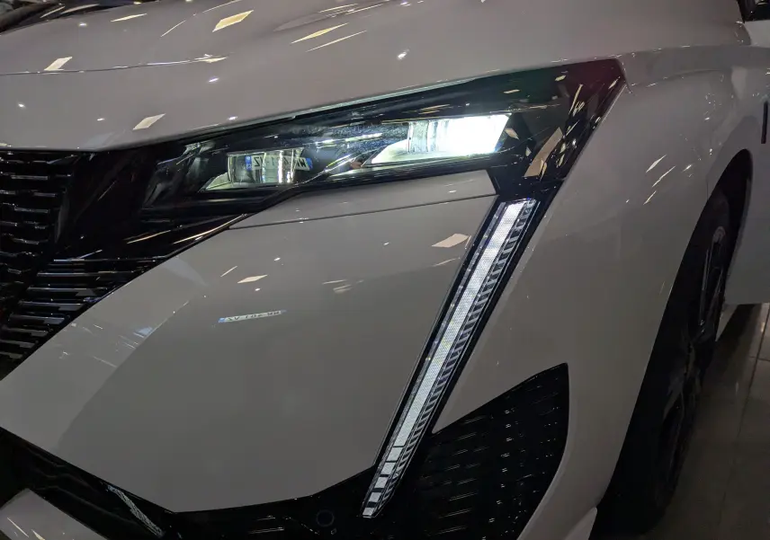 Gros plan sur le phare avant droit allumé d'une Peugeot 308 GT blanche Okénite avec signature lumineuse verticale LED.