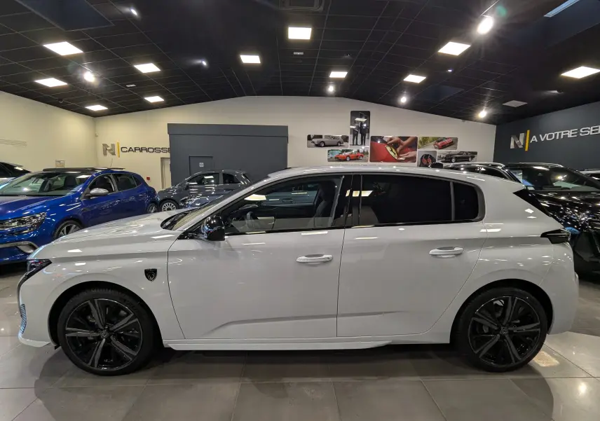 Profil côté gauche de la Peugeot 308 GT blanche Okénite 2024 avec jantes noires et vitres teintées en showroom.