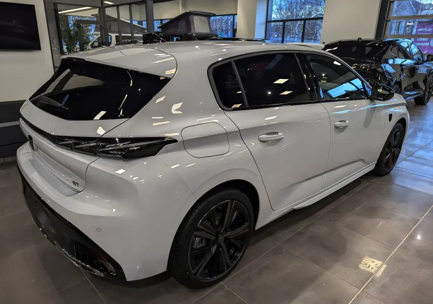 Vue 3/4 arrière droite d'une Peugeot 308 GT 2024 blanc Okénite avec jantes noires et vitres teintées en showroom.