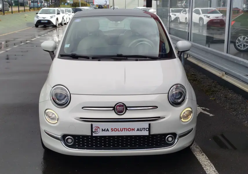 Vue frontale d'une Fiat 500 blanche Bossa Nova, avec ses phares ronds caractéristiques allumés, stationnée devant un bâtiment vitré.