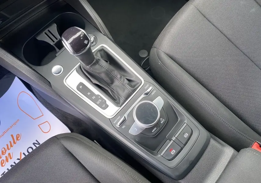 Vue rapprochée de la console centrale noire de l'Audi Q2 2023, avec levier de vitesse automatique et molette de contrôle multimédia.
