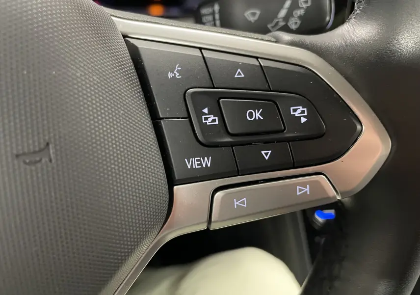 Gros plan sur les commandes au volant côté gauche du Volkswagen Touran gris Dauphin 2024, avec boutons multifonctions éclairés.