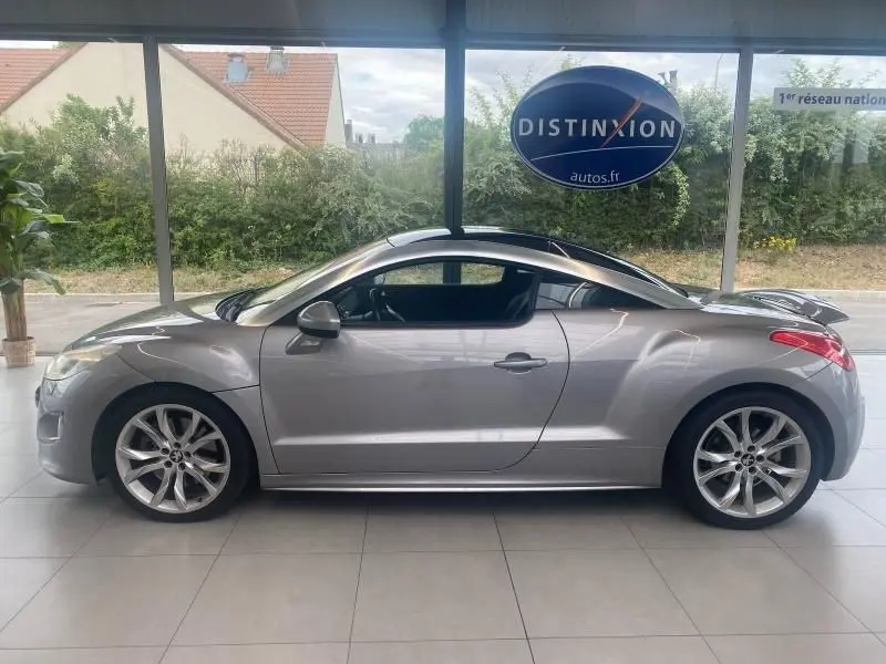 Peugeot RCZ gris vu de profil côté gauche, coupé sportif avec jantes alliage et toit double bossage visible en intérieur showroom.