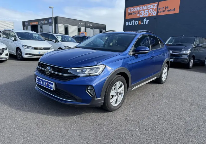 Volkswagen Taigo bleu récif métallisé vu en 3/4 avant droit sur parking devant un panneau publicitaire orange.