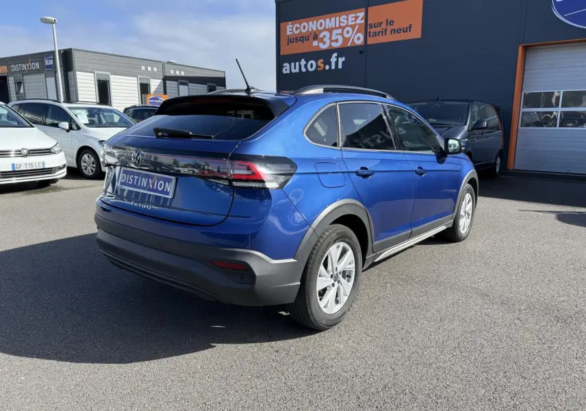 Volkswagen Taigo bleu récif métallisé vu en 3/4 arrière droit sur parking devant un bâtiment commercial.